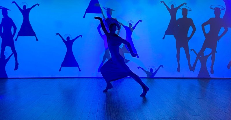 A dancer in a light dress poses center-frame, silhouetted against a bright blue wall with multiple dark blue projected figures.