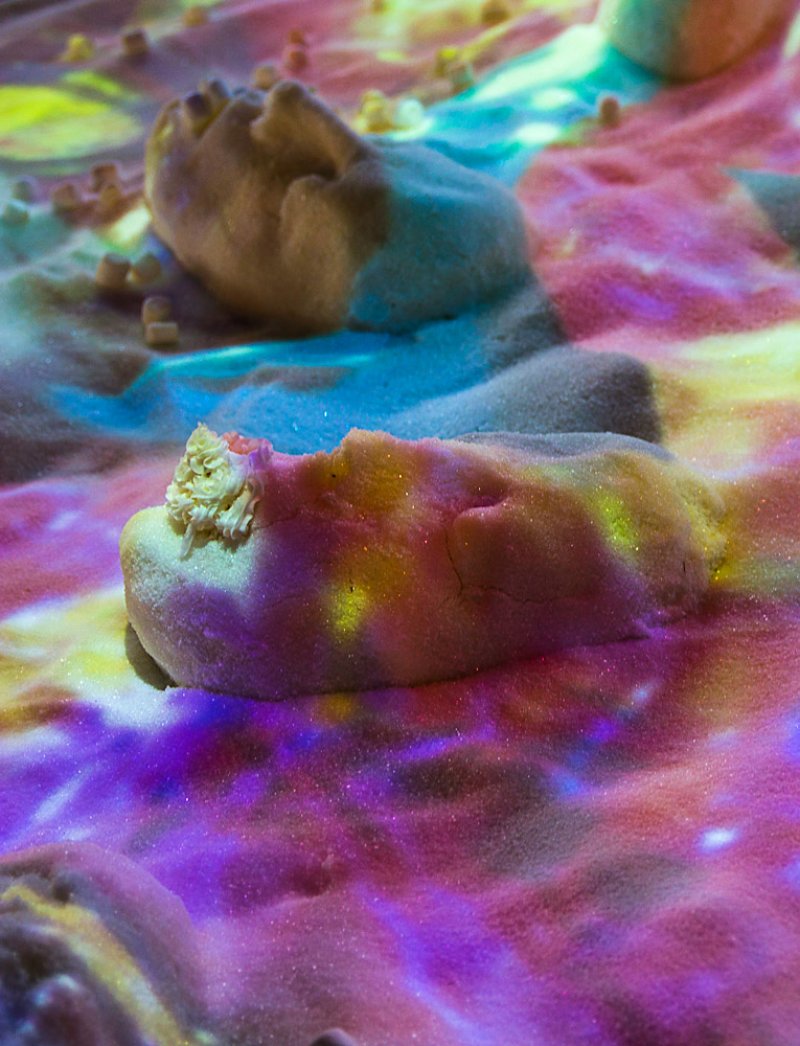 Close-up of a sand installation with abstract forms, illuminated by shifting pink, purple, yellow, and blue light.