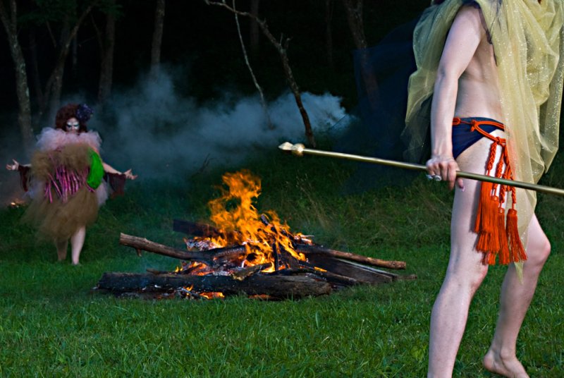 Two figures in elaborate costumes by a bonfire in a dark forest. One holds a staff, the other stands in smoke.