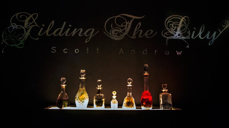 Seven glass decanters with stoppers, filled with liquids of varying colors, sit on a shelf against a dark background. Above, the title "Gilding The Lily" and artist's name, Scott Andrew, are displayed in a decorative font.