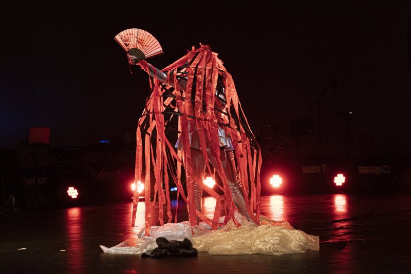 Performance art piece with a figure in silver bodysuit, partially obscured by a cage-like structure draped with red ribbons, holding a fan. Stage lighting creates a dramatic, dark atmosphere.