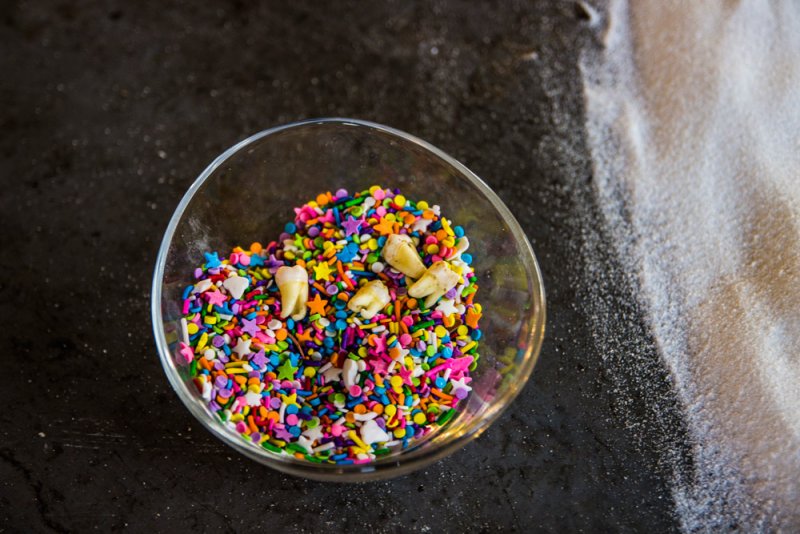 Glass bowl filled with colorful sprinkles and four human teeth, next to a pile of white powder.