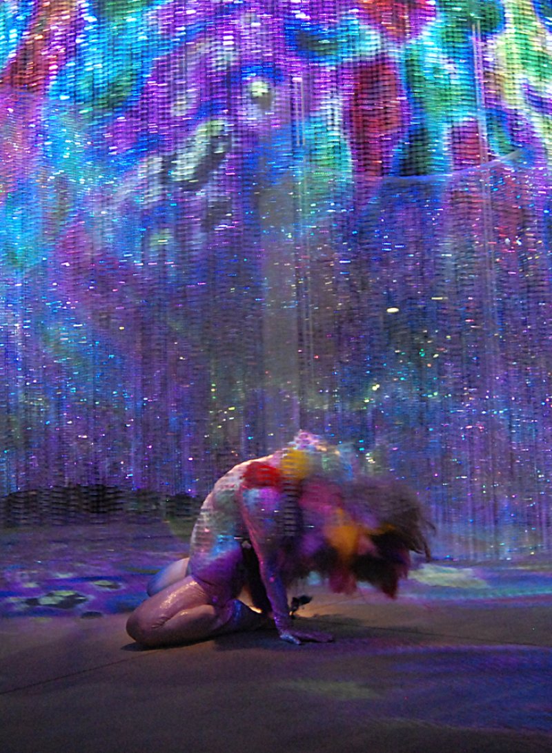 A dancer in a dark space, illuminated by vibrant blue, purple, and pink light from above, bending over with hair in motion.