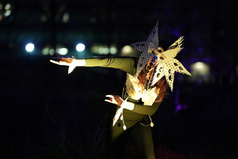 Performer in a dark setting, wearing a star-shaped, reflective headpiece with circular cutouts, arms outstretched.