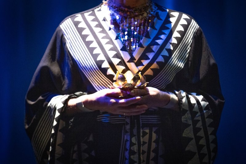 Person in patterned black and white robe holding a golden lotus, with a shimmering beaded necklace, against a dark blue background.