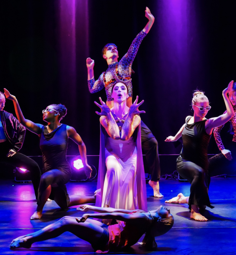 Seven dancers in a dramatic pose on a stage with purple and blue lighting.