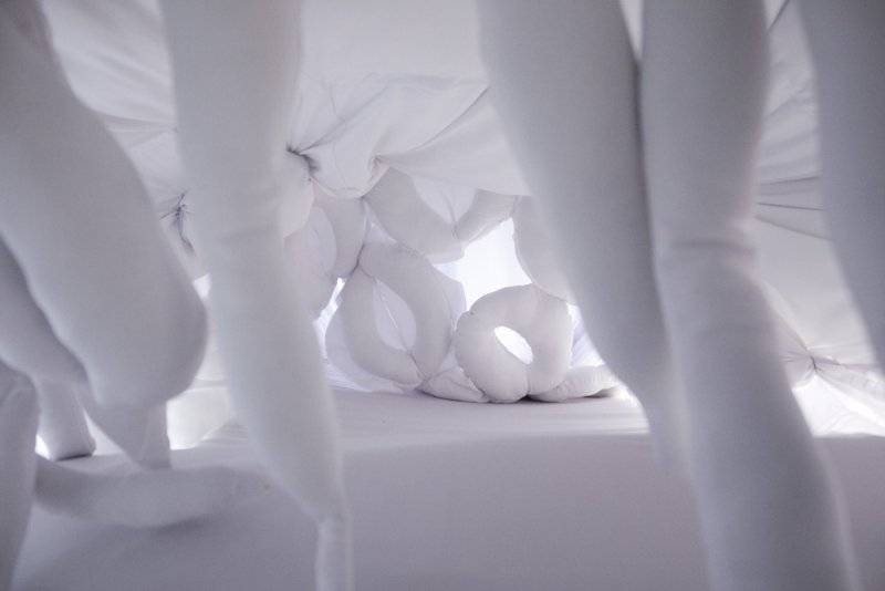 Soft white fabric sculpture with dangling limbs and round, pillow-like forms, creating a cocoon-like space.