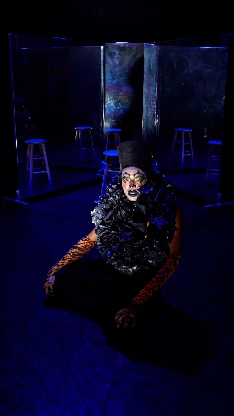 Performer in black costume, white face paint, and tiger-striped gloves sits on a dark stage with stools and reflective panels.