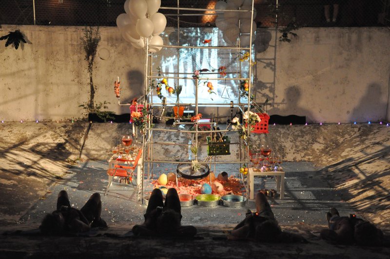 Installation with white balloons, drinks, and objects on a metal structure, projecting a forest scene onto a concrete wall.