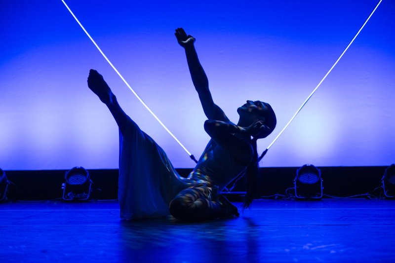Dancer in flowing dress, lit in blue, poses with arms raised against a bright backdrop with angled light bars.