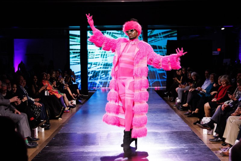 A person in a vibrant pink furry coat, matching pants, hat, and sunglasses poses on a runway with an audience seated on both sides.