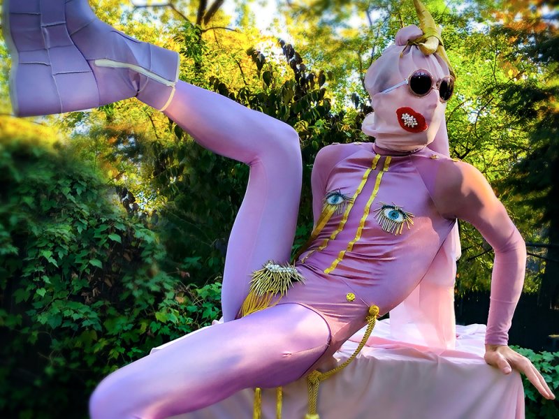 Performance art featuring a person in a shiny pink bodysuit, mask, sunglasses, and unicorn horn, with gold accents.