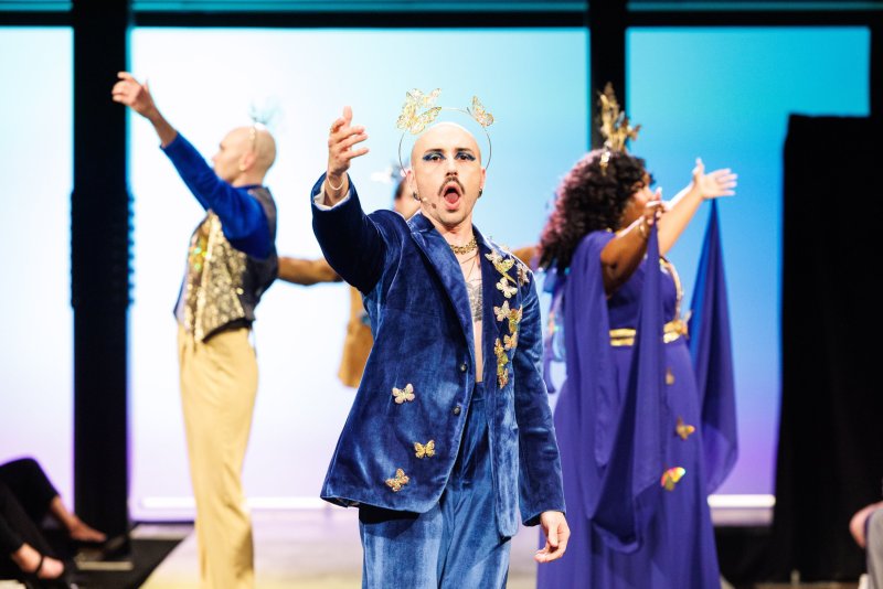 Performer in blue velvet suit with gold butterflies and butterfly crown, singing center stage, with two other performers behind.