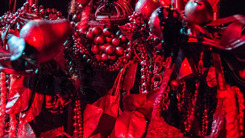 Close-up of a dense, dark red sculpture with beaded chains, leaves, and spherical elements under dramatic red lighting.