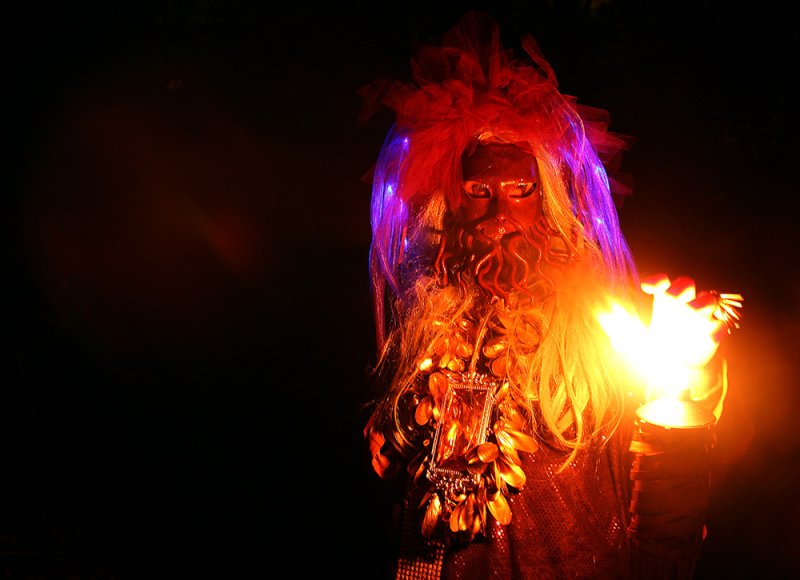 Figure in a dark setting with a gold mask, long blonde hair, and a red headdress, holding a flaming torch.