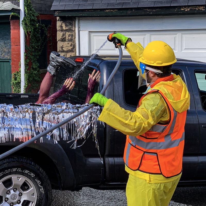 Man in yellow hazmat suit sprays water into a truck bed with legs and arms emerging from silver tinsel.