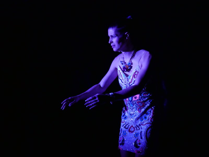 Woman in a short, patterned dress and large necklace stands in near darkness, illuminated by purple light. She faces left with her hands outstretched.