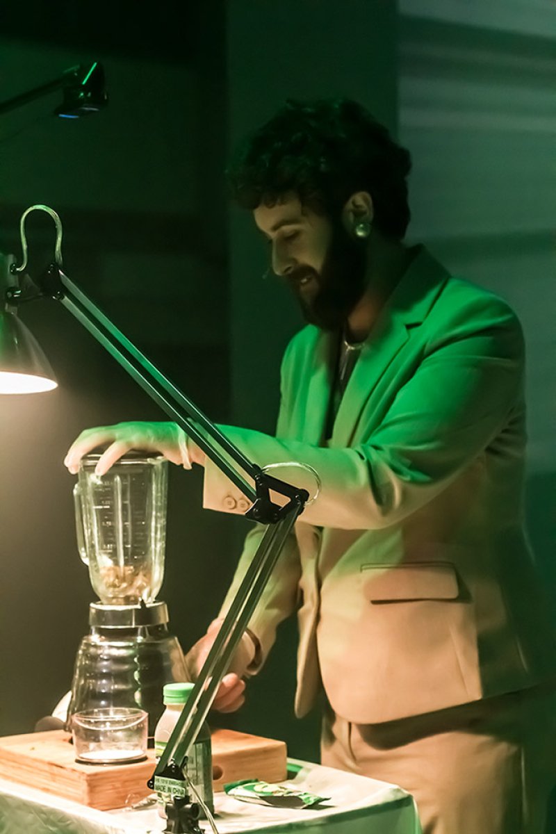 Man in a light green suit jacket operating a blender on a table, bathed in green light.