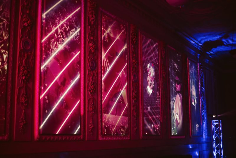 Digital art installation with neon pink and white diagonal lines, and figures in formal wear, projected on ornate mirrored panels.