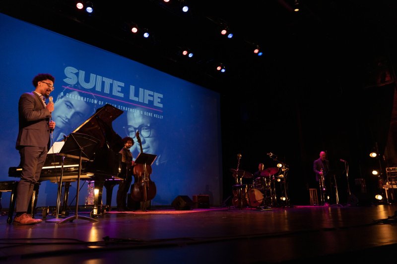 A jazz band performs on stage with a large blue screen displaying "SUITE LIFE" and portraits of musicians.