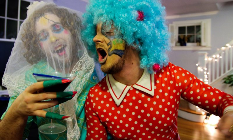 Two men in drag, one with a blue wig and polka dot dress, the other with a veil and painted face, holding a martini.