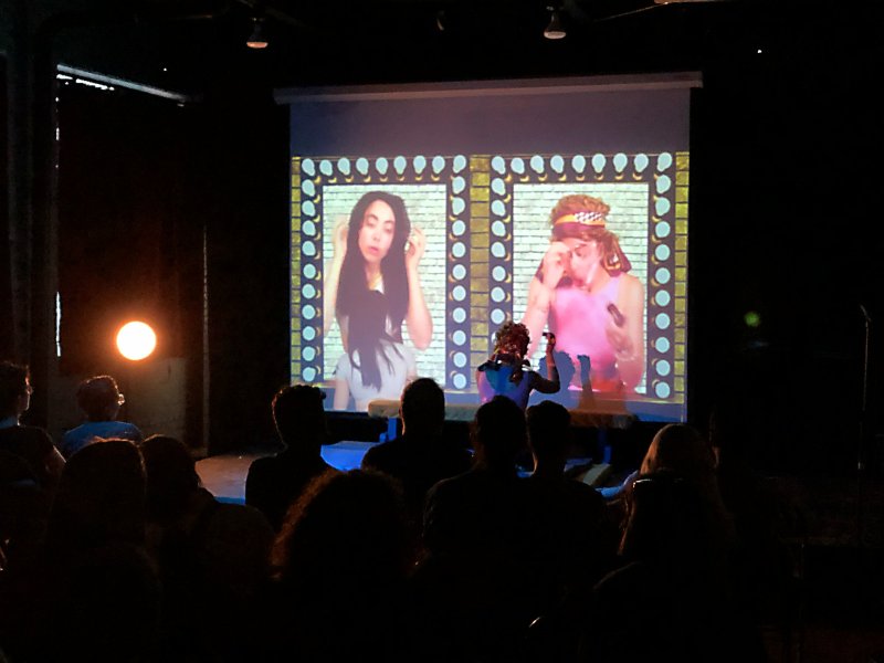 Dark room with silhouetted audience watching a projection of two portraits framed by brick and lightbulb patterns. The portraits feature two figures in retro attire.
