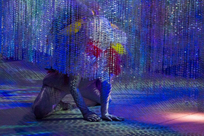 Performance art with a figure in a shimmering costume kneeling amidst hanging crystal strands and blue light projections.