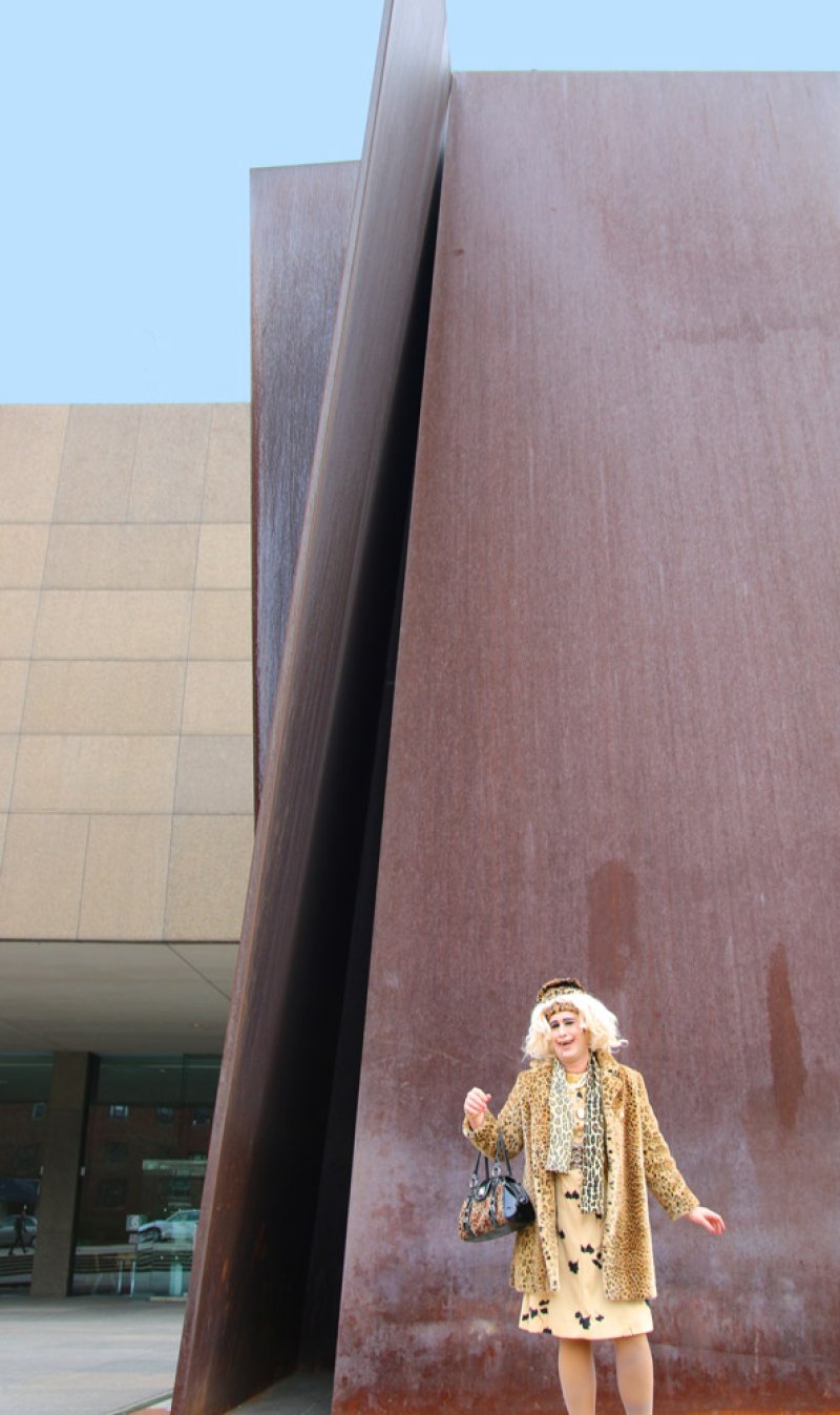 Blonde woman in leopard print coat, dress, and headband, holding a purse, stands before a large rusted metal sculpture.