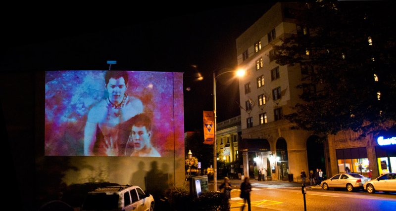 Nighttime outdoor projection of two shirtless men with blue and pink hues on a building, cars and people below.