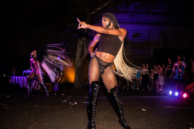 Performance shot with two drag queens in black crop tops, sparkly fishnets, and high black boots. One figure points, the other has long hair flowing. Audience visible in background. Purple lighting.