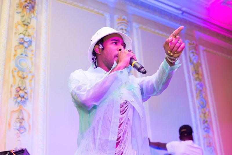 Performer in white bucket hat and sheer shirt sings into a microphone, pointing. Stage lighting casts blue and pink hues. Ornate architectural details in the background.