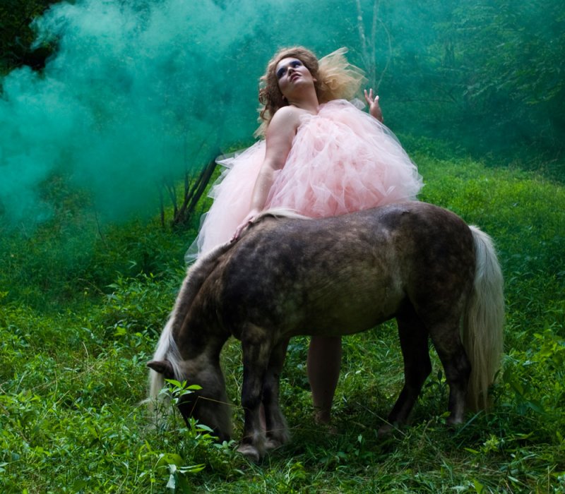 A person in a voluminous pink gown sits atop a brown pony grazing in a lush green forest, with green smoke swirling.