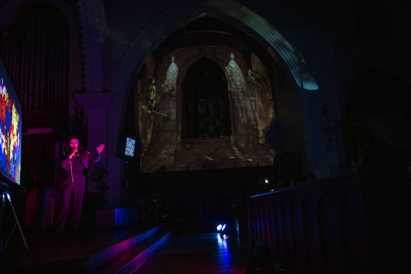 Performer in dark jumpsuit on stage, singing into a microphone, with colorful digital art and a projected gothic archway.