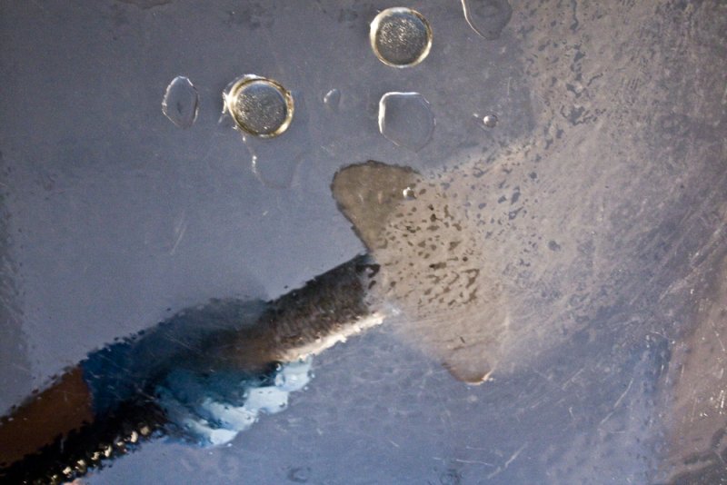 Close-up of a hand holding a dark, textured object spraying water onto a scratched, light gray surface with water droplets.