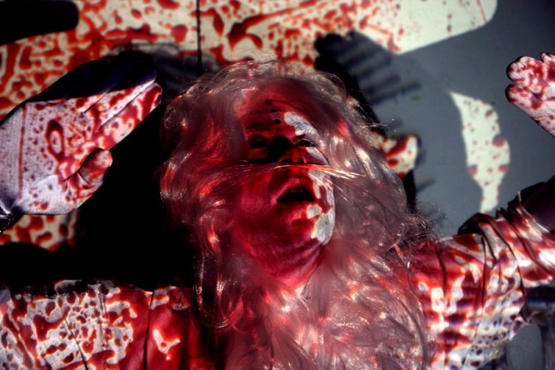 A person with white hair and face paint, covered in projected red blood-like patterns, hands raised.