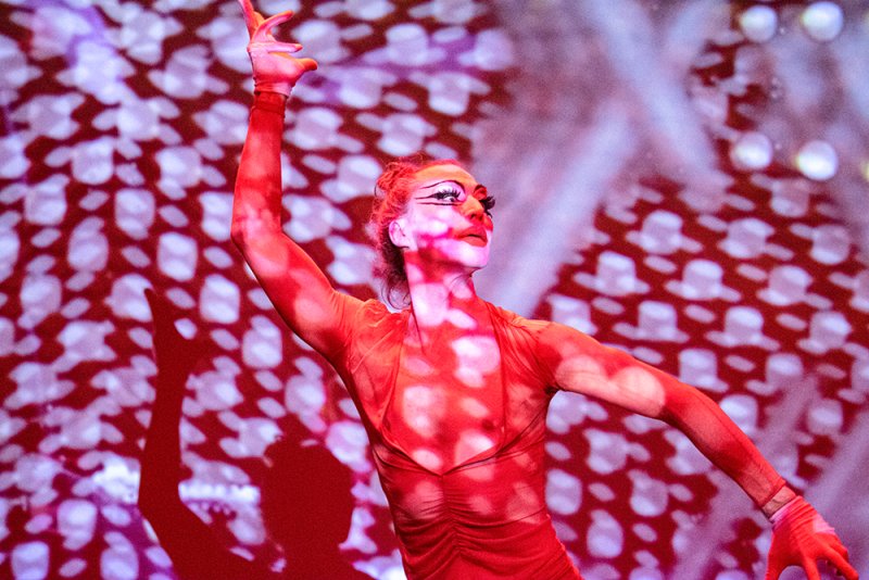 Performer in red, with dramatic makeup and arm raised, against a projected red and white patterned background.