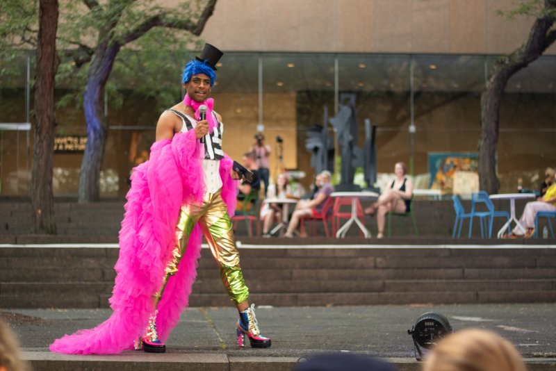 Performer in a black top hat and blue hair, wearing a pink tulle boa and gold pants, stands on a stage holding a microphone. People sit at tables in the background.