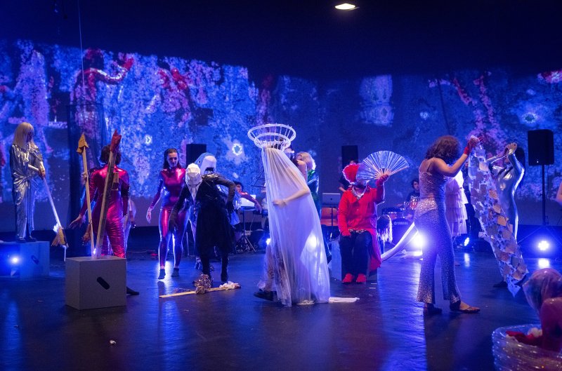 Performance art with costumed figures in a dark room with blue and purple projected abstract visuals on the back wall.