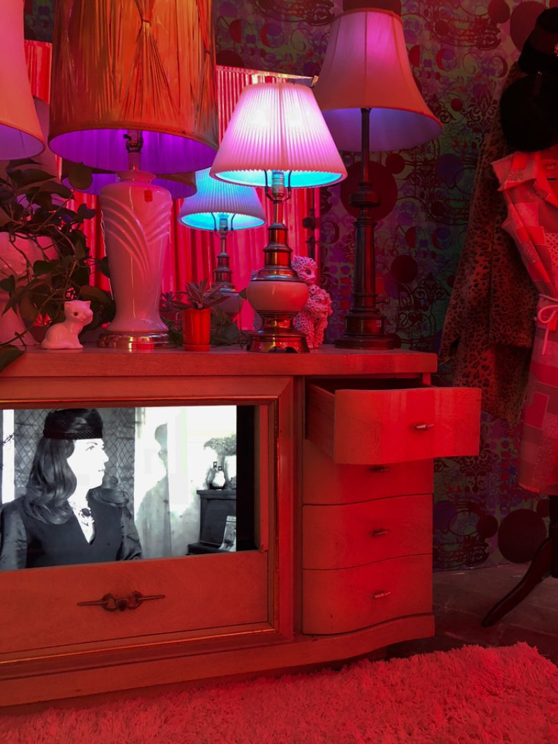 A vintage dresser with a black and white video playing on its mirror, surrounded by brightly lit lamps and colorful decor.