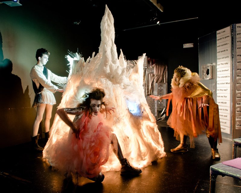 Performance art with three figures in elaborate costumes around a glowing, jagged white structure.