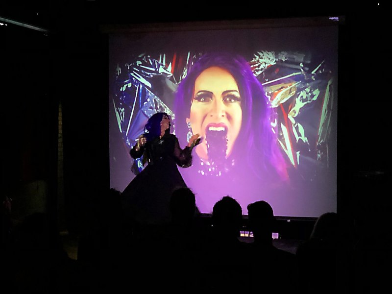 Performance art: a drag artist in black dress stands before a projection of a face with purple hair and open mouth.