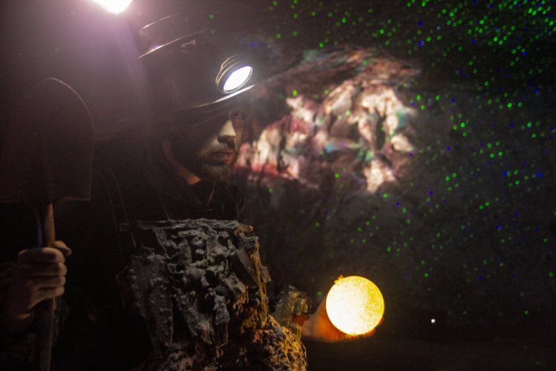 A person in mining gear holding a shovel and a glowing yellow orb, with green laser dots in a dark cave.