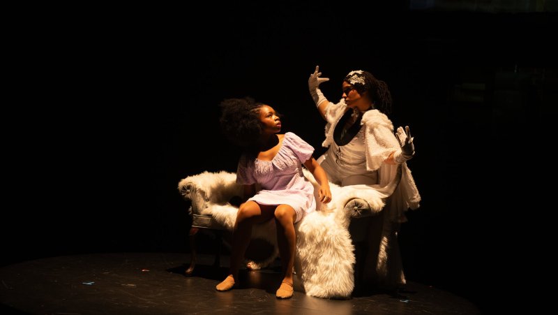 Two figures on a stage, lit by a spotlight. One in a lilac dress sits on a fur-covered chair. The other, in white gloves and dress, stands behind with an ornate headpiece. Dark backdrop.