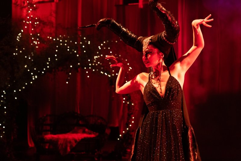 Woman in a black sequined dress and elaborate horned headpiece, dancing under red lights with sparkling fairy lights in the background.