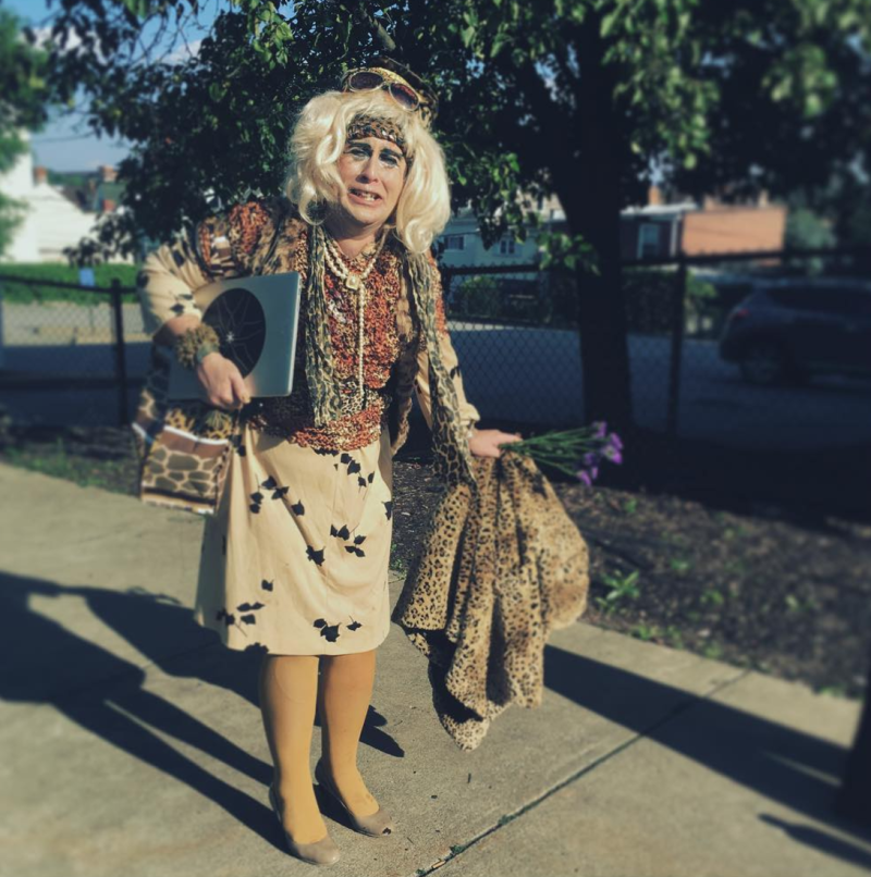Blonde person in drag wearing leopard print, a tan skirt, and yellow tights, holding a laptop and flowers outdoors.