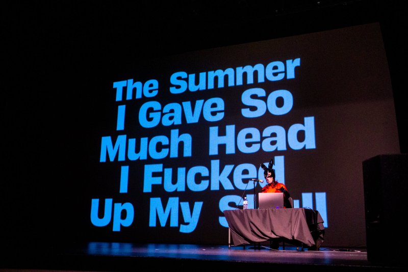 Performance art with a person in a bunny mask and orange outfit at a table with a laptop. Behind them, a large screen displays blue text on a black background.