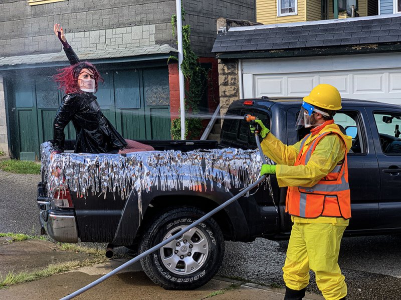 Outdoor scene with a person in a yellow hazmat suit spraying water on a person with red hair sitting in the back of a pickup truck decorated with silver tinsel. Both wear face shields.