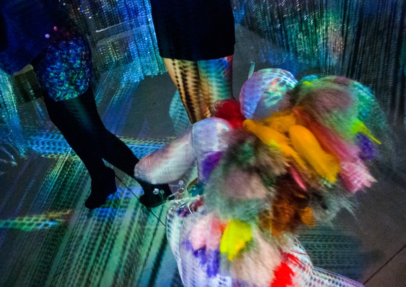 Two people's legs in a vibrant, abstract light installation with colorful, furry sculptures on the floor.