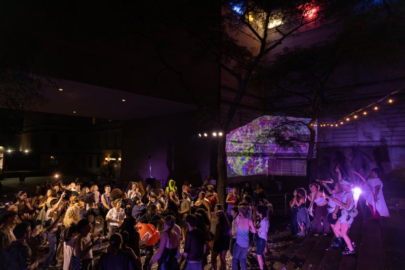 A nighttime outdoor gathering with a diverse crowd dancing under colorful lights, a large tree, and a building with projections.
