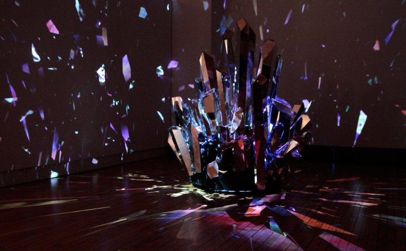 Sculpture of mirrored crystal shards reflecting purple and blue light onto a dark room and wooden floor.